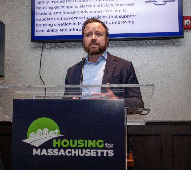 A man wearing a brown blazer and blue buttondown shirt stands speaking at a podium that has a sign on it saying Housing for Massachusetts. Behind him is a screen with out of focus words.